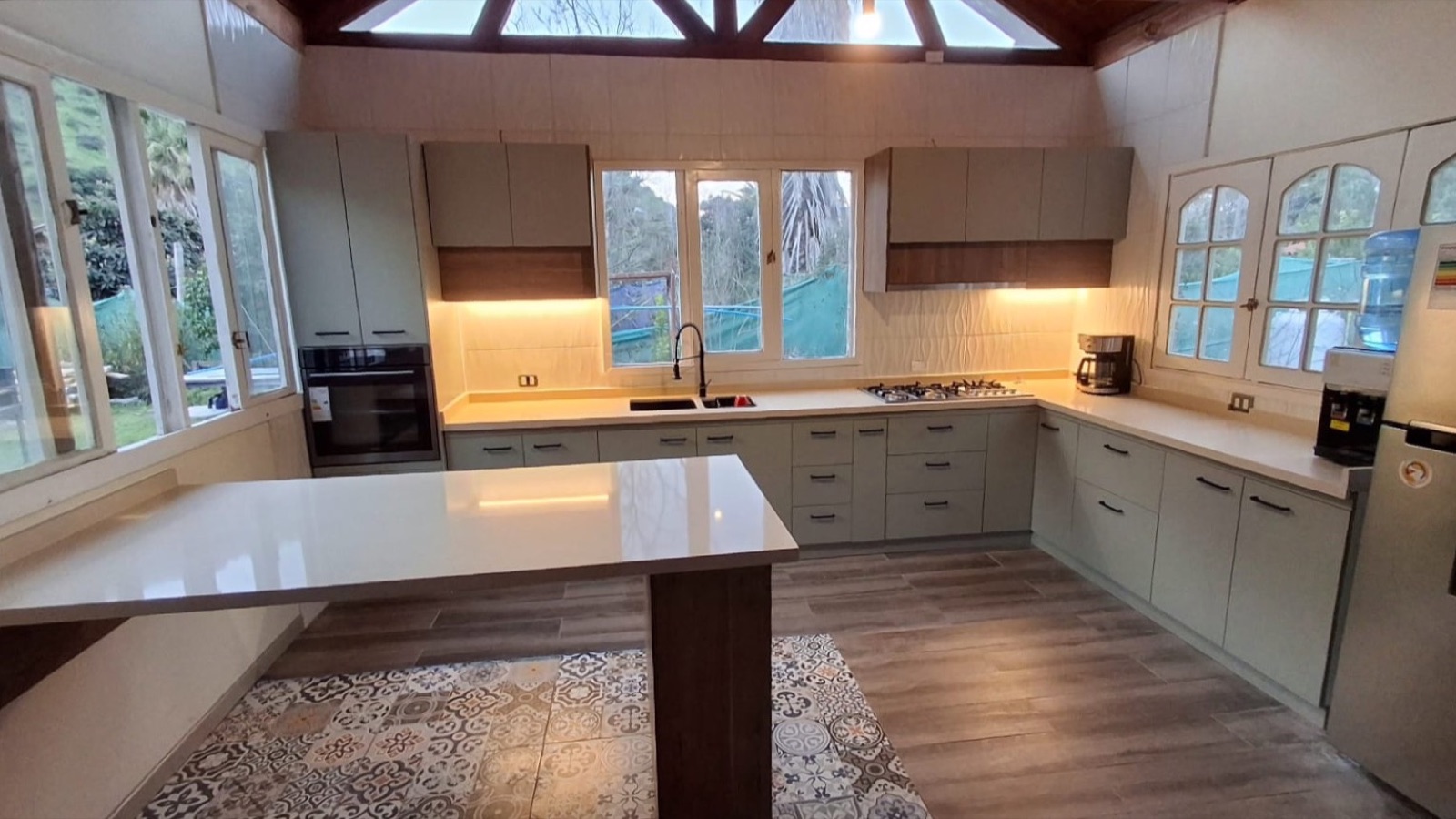 Cocina en U color arena con isla central, vigas de madera a la vista y ventanales al bosque