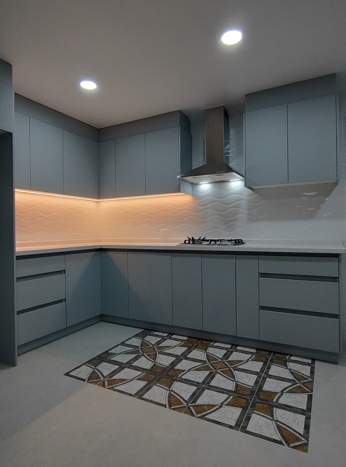 Cocina en L gris azulado con campana de acero, luz LED bajo los muebles altos y piso decorado