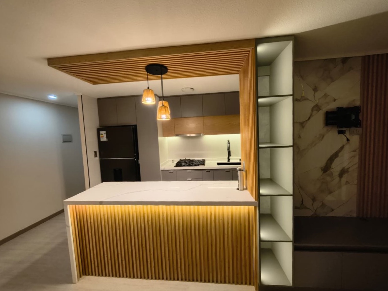 Isla de cocina con frente de listones de madera iluminado, cubierta de mármol y lámparas colgantes