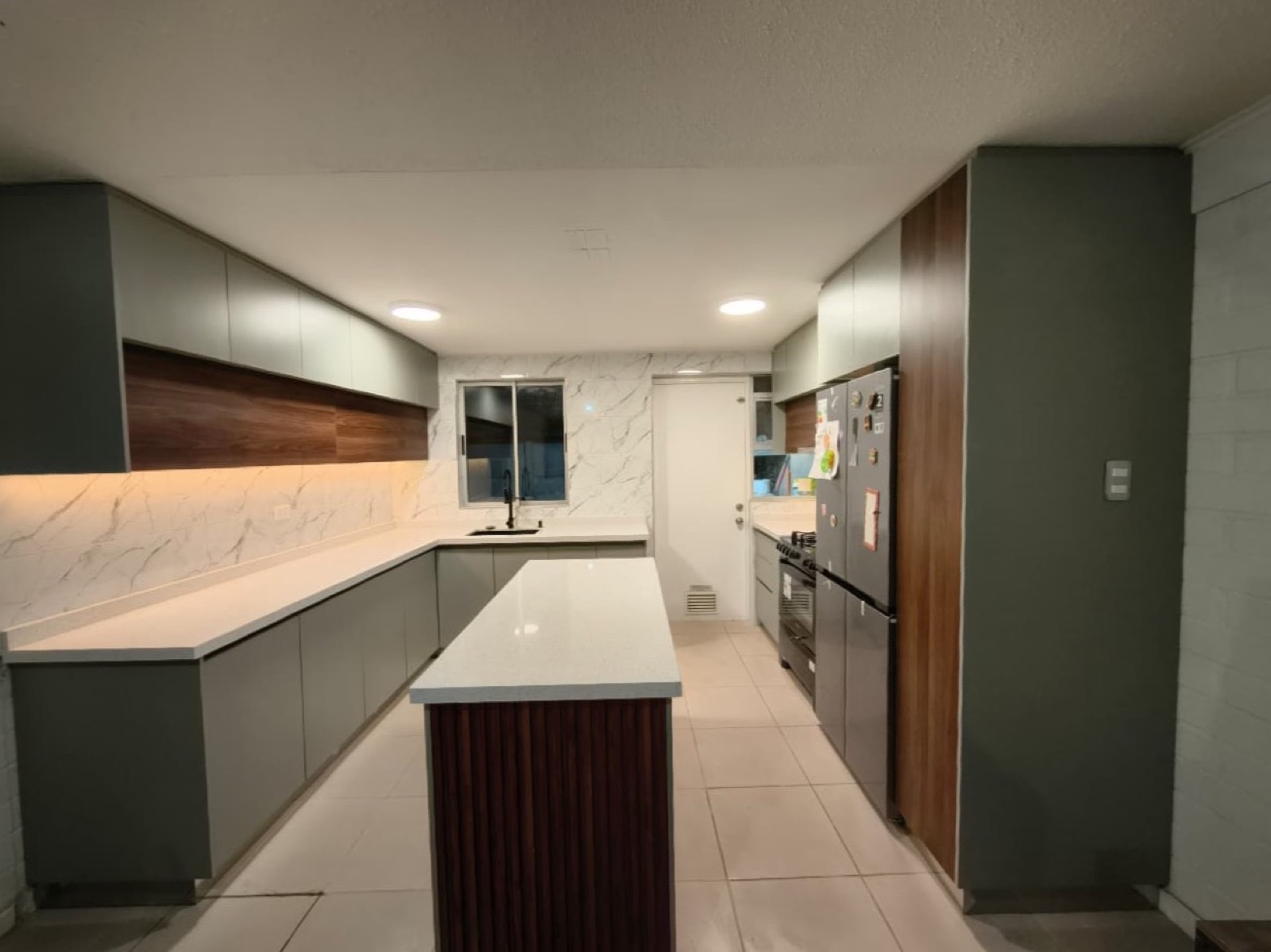 Cocina verde salvia con isla, muro de mármol y despensa alta de madera