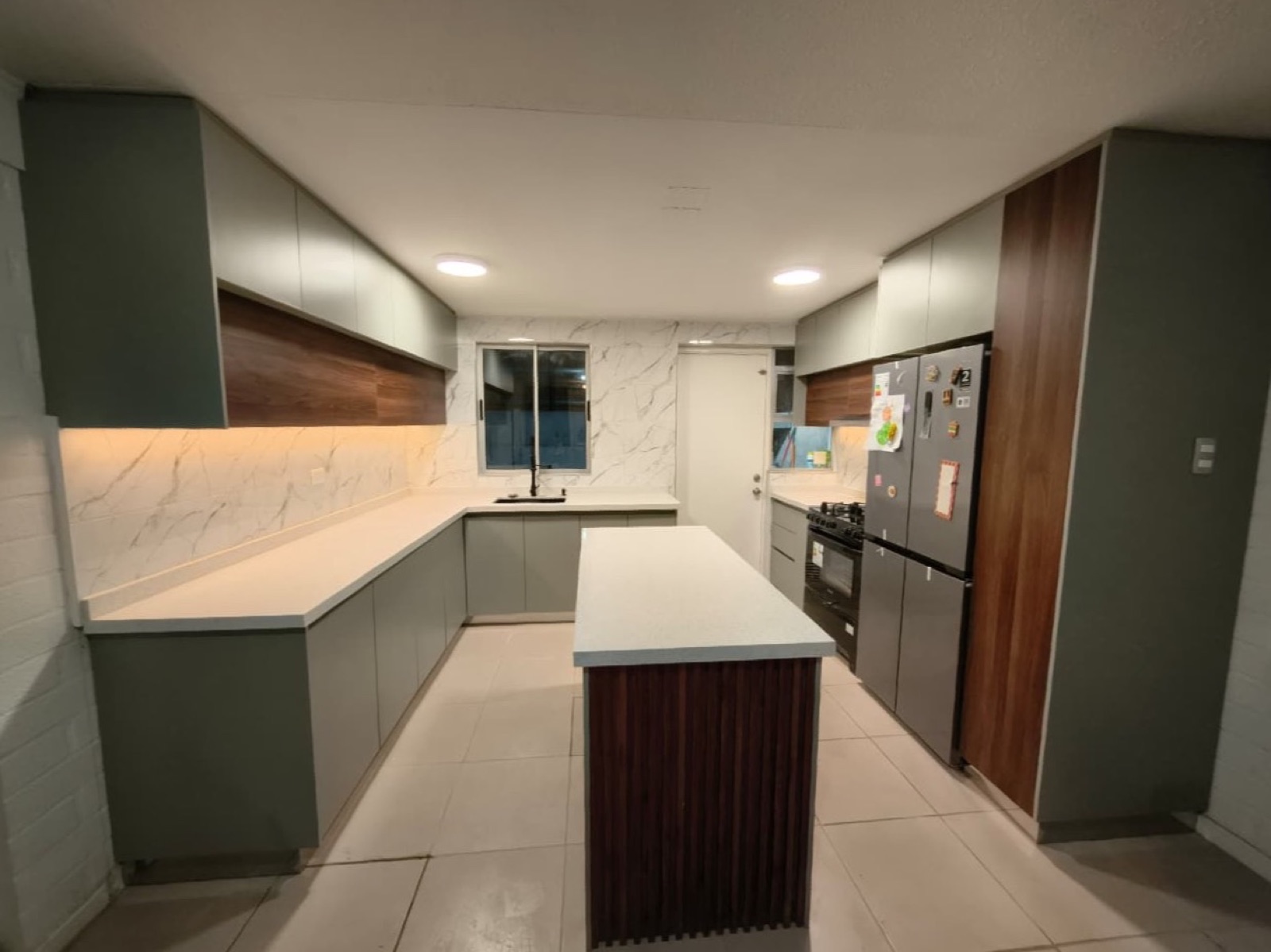 Cocina verde oliva con isla de madera, muro de mármol y refrigerador empotrado