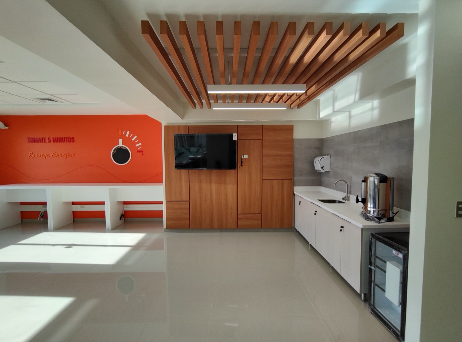 Comedor de personal con kitchenette, mesón corrido con puestos individuales, mueble mural para televisor y cielo decorativo de listones de madera.