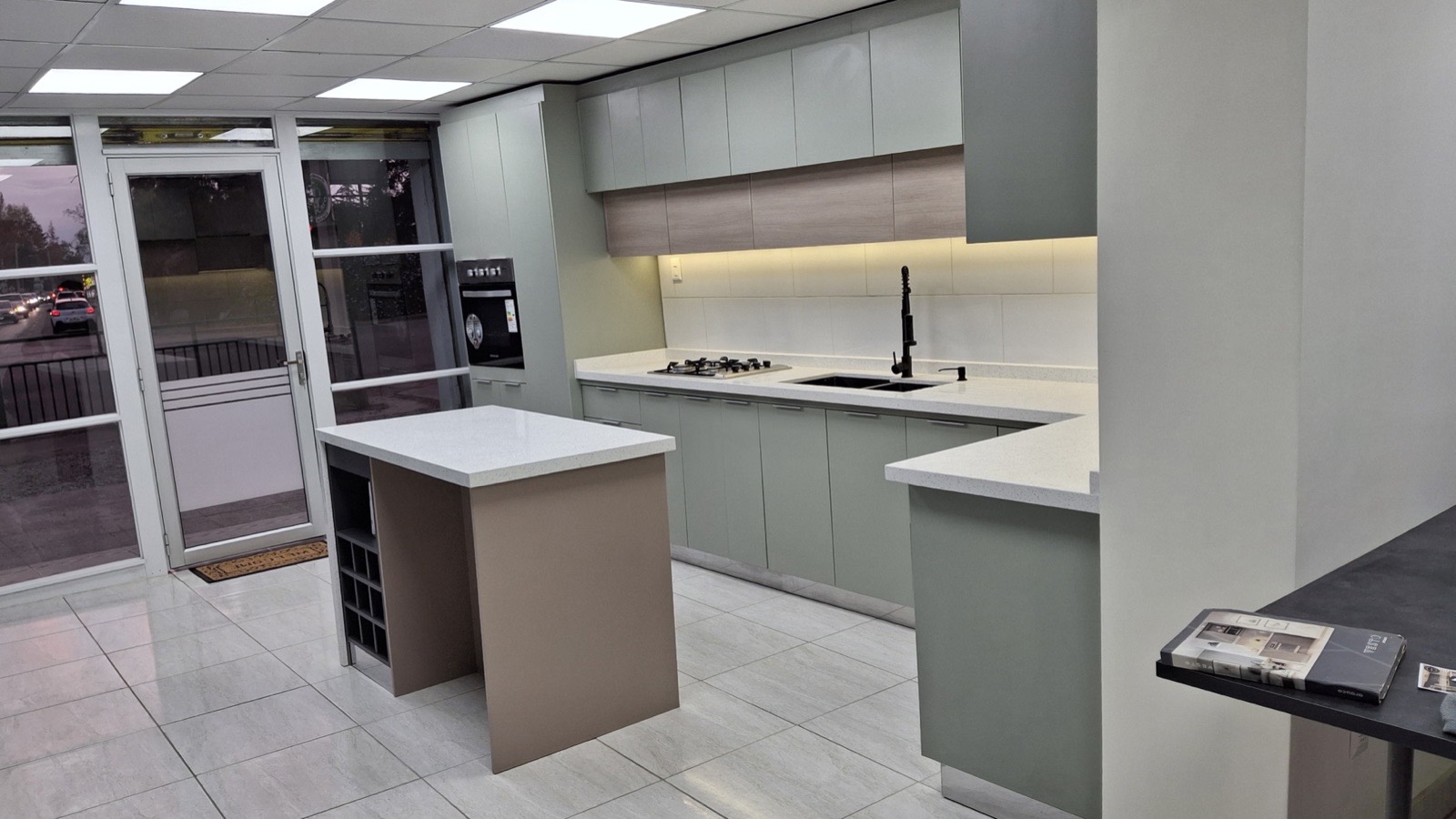 Showroom con muebles de cocina, columna horno y despensa en melamina verde glaciar. Isla pata cascada, con botellero y cubiertas en cuarzo blanco galaxy.
